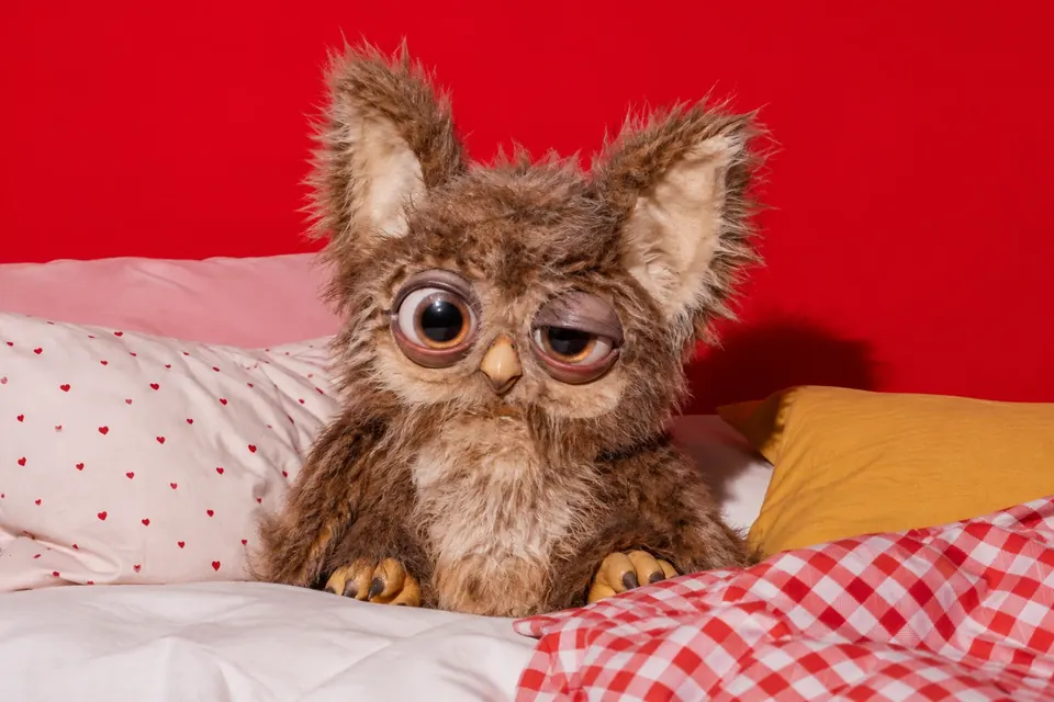 Ein flauschiges, eulenartiges Plüschwesen mit riesigen Augen und großen Ohren sitzt auf einem Kinderbett