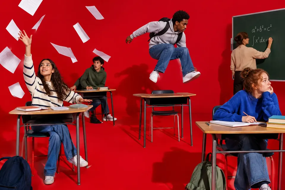 Eine Schulklasse ist mitten in der Bewegung erstarrt, Papiere hängen in der Luft, ein Schüler springt über einen Tisch